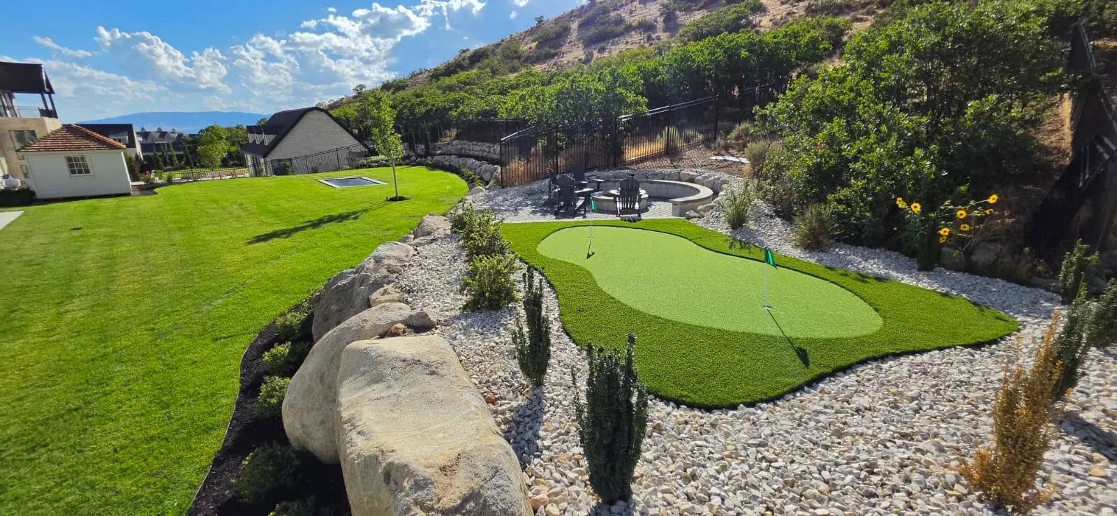 Putting green bordered by gravel and boulders beside a lawn and circular fire pit patio