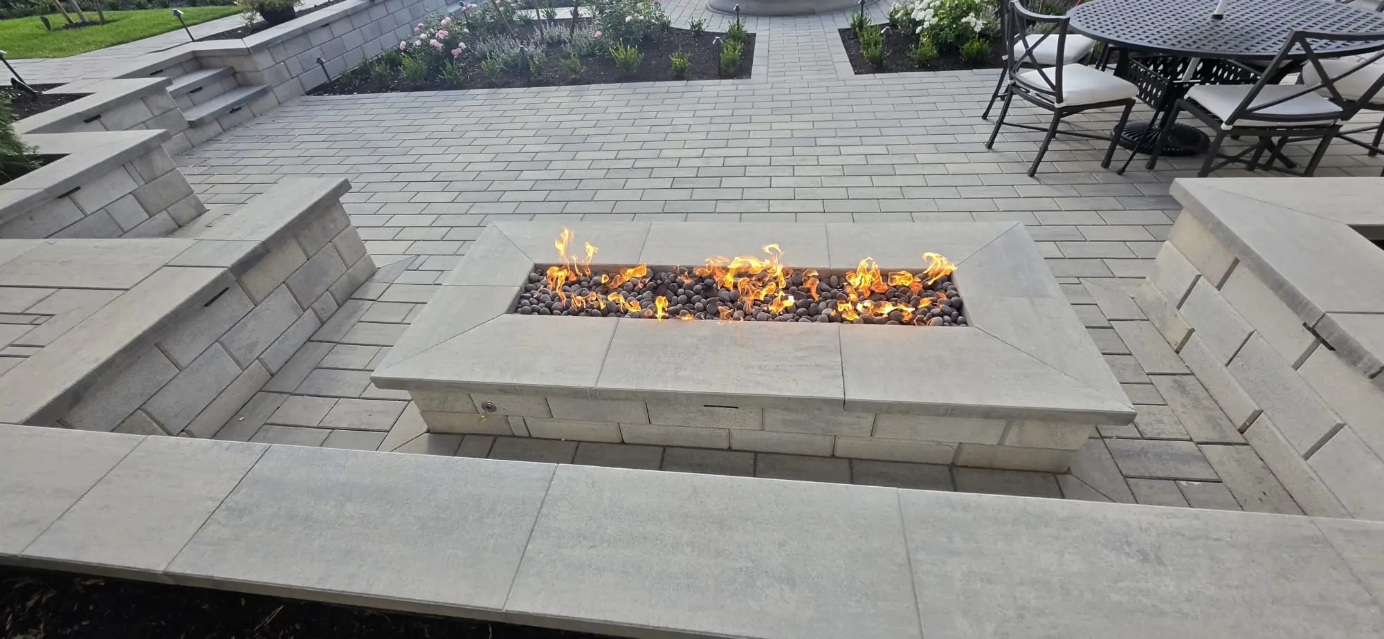 Rectangular fire pit with flames beside a paver patio and built-in seating