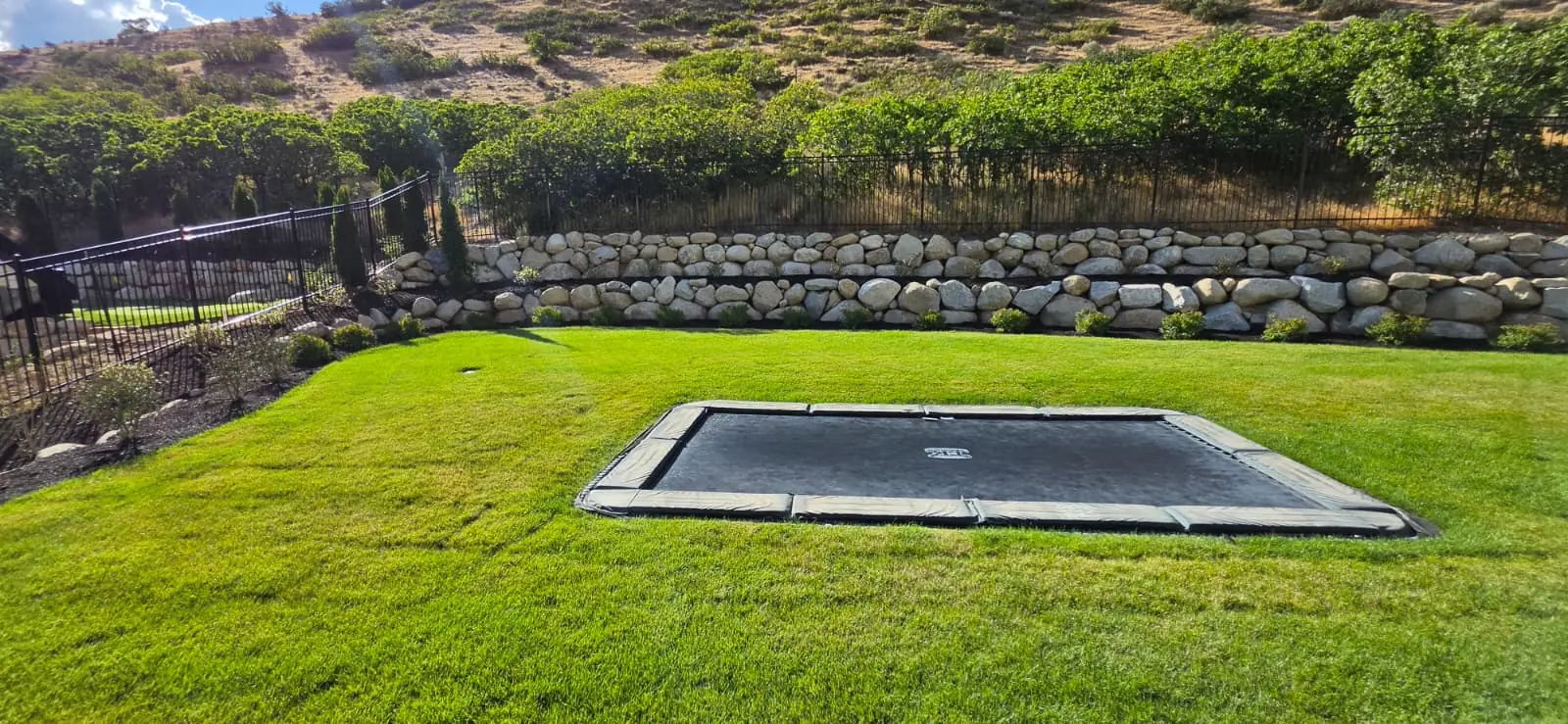 Rectangular in-ground trampoline surrounded by lawn and a boulder retaining wall